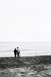 A black-and-white image captures two people walking on a beach, facing away from the camera. Holding hands, they stroll by the ocean with calm waves. The sky and sea blend together in "Errance à Ostende," creating a serene and peaceful atmosphere.